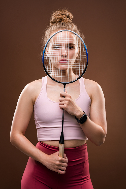 Young pretty female tennis player in pink tanktop and crimson leggins holding racket in front of her face while standing in isolation
