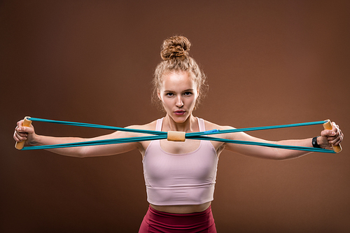 Young active female in tracksuit stretching elastic band in front of herself and looking at you while exercising in front of camera in isolation