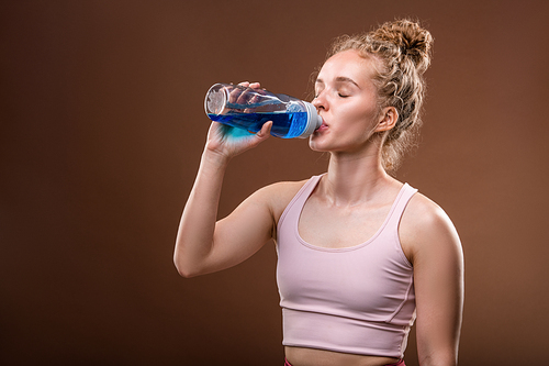 Young fit thirsty sportswoman drinking water from plastic bottle while having short break for rest and refreshment after sports training