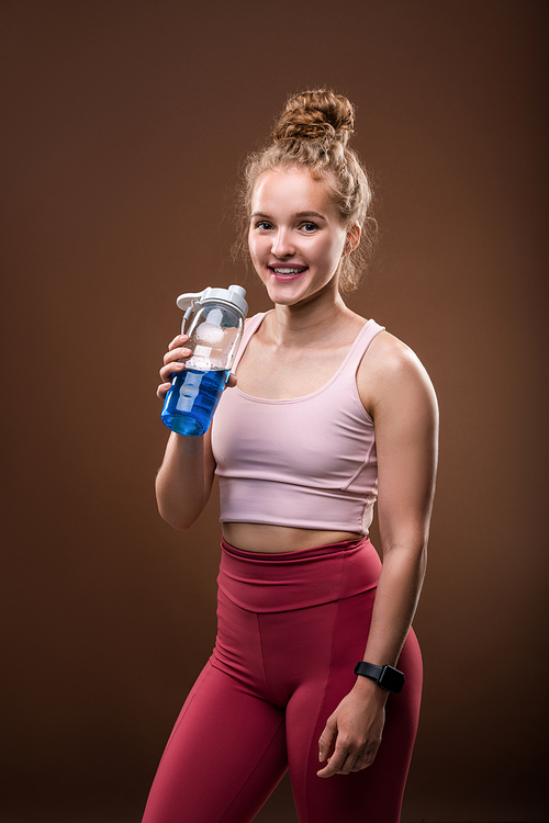 Young cheerful sportswoman with toothy smile holding plastic bottle with refreshing drink while having break after training in gym