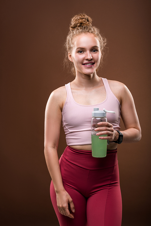 Young fit woman in tracksuit holding bottle with drink while having short break after sports training against brown background in isolation