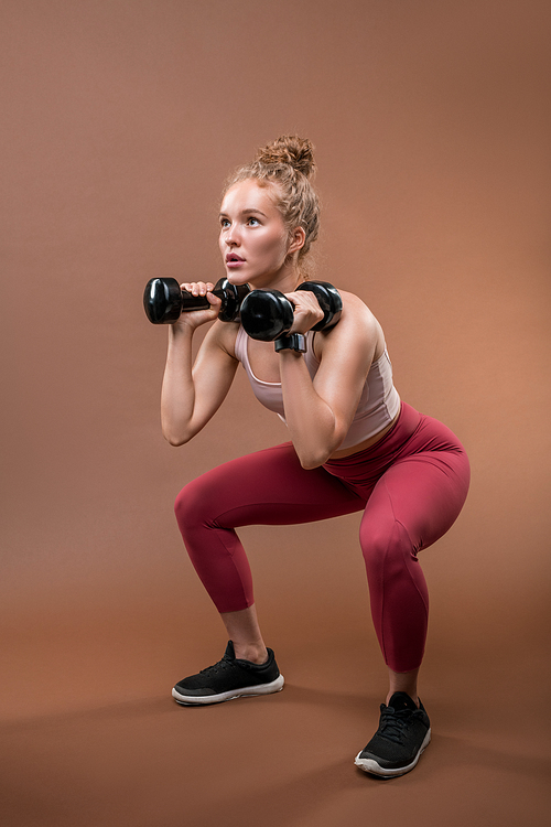 Young strong fit sportswoman in tracksuit sitting on squats and lifting dumbbells while making effort during difficult exercise