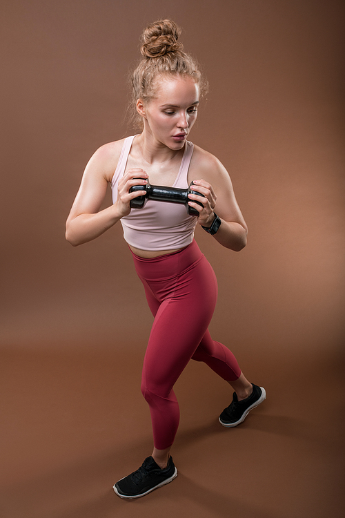 Active young blond woman in sportswear holding dumbbell with her arms bent in elbows during morning workout against brown background