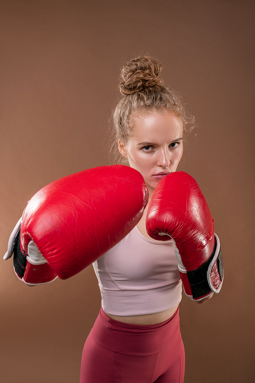 Young serious active woman in tracksuit and red boxing gloves doing kicks and hits in front of camera during individual training in isolation