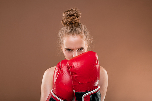 Young aggressive female athlete with blond curly hair holding hands in red boxing gloves close to her face while looking at you before fight