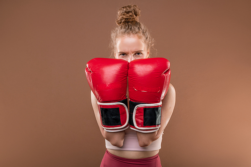 Young blond sportswoman in tracksuit keeping hands in boxing gloves by her face while standing in front of camera during individual training
