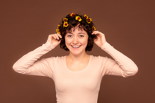 Joyful young brunette female of Caucasian ethnicity with small flowers in dark short curly hair touching her head while standing in isolation