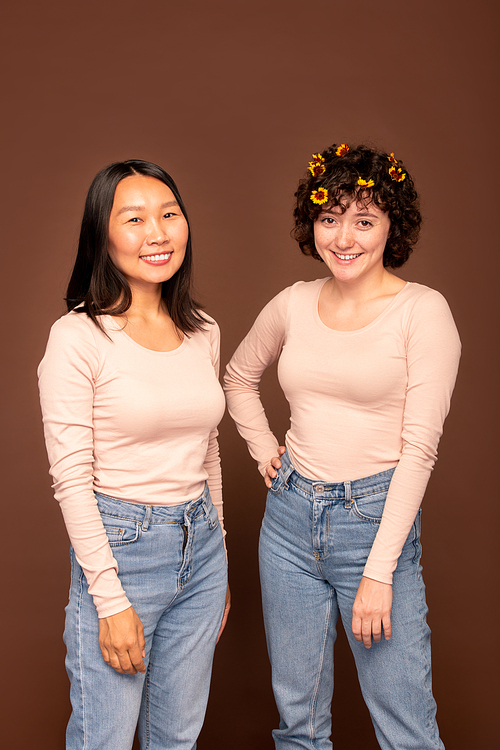 Two young happy intercultural women in white pullovers and blue jeans standing close to one another in front of camera and looking at you