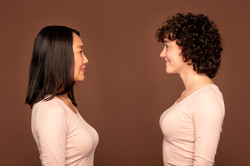 Side view of two cheerful young females of Asian and Caucasian ethnicities in white pullovers looking at each other over brown background