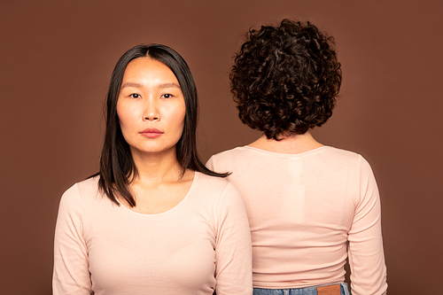 Young serious brunette woman of Asian ethnicity in white pullover standing in front of camera on background of back of another female