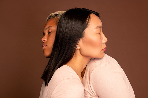Young Asian and African-american females with their eyes closed embracing one another and keeping heads on shoulders of each other