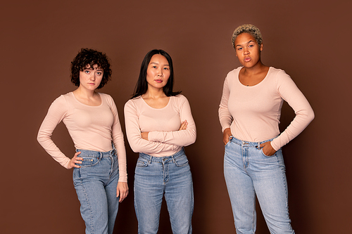 Three young intercultural females in white pullovers and blue jeans standing in row against brown background and looking at you