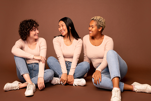 Group of young casual females of various ethnicities sitting on the floor against brown background, looking at each other and chatting