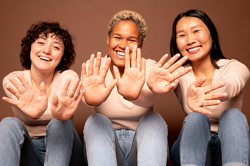 Joyful young smiling women of Asian, Caucasian and African ethnicities showing their palms to you while sitting in row in front of camera