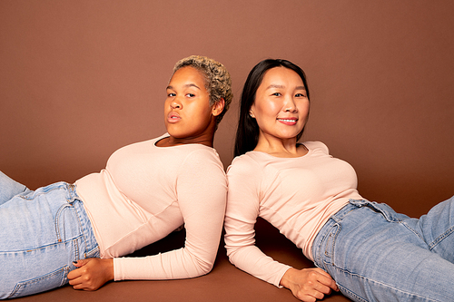 Two young contemporary females of various ethnicities in blue jeans and white pullovers lying on the floor by wall in front of camera