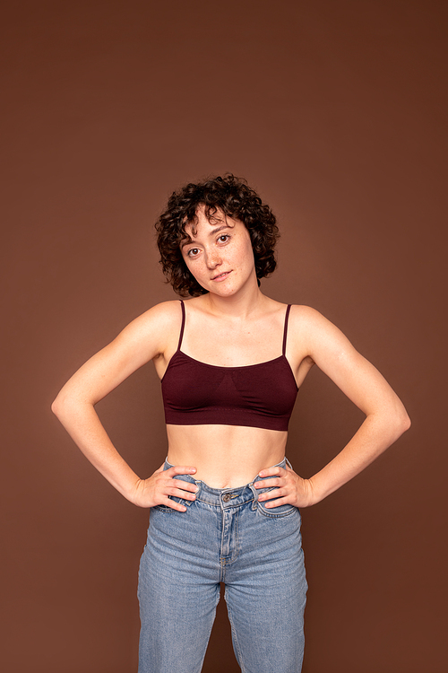 Young gorgeous Caucasian brunette woman with short curly hair keeping her hands on waist while standing over brown background in isolation