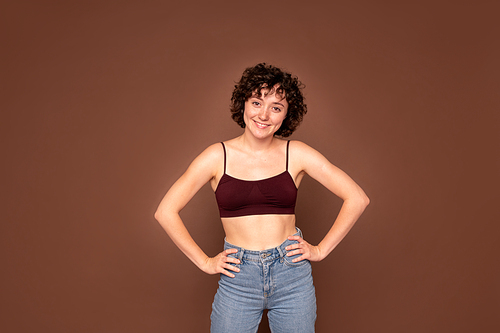 Happy young gorgeous Caucasian woman with short dark curly hair keeping her hands on waist while standing by wall in isolation