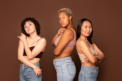 Young beautiful intercultural females in tanktops and blue jeans standing in row against brown background in front of camera and looking at you