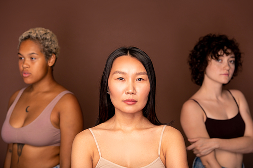 Young serious Asian woman in beige tanktop standing in front of camera against two other females of various ethnicities and looking at you