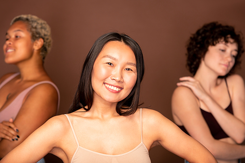 Young cheerful Asian woman in beige tanktop standing in front of two other intercultural females and looking at you with toothy smile
