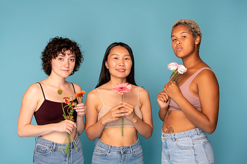 Three young pretty women in tanktops and blue jeans holding fresh flowers while standing against blue background in front of camera