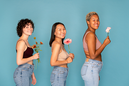Three young cheerful women in tanktops and blue jeans holding fresh flowers in front of themselves while standing in front of camera