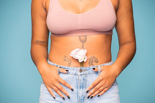 Low section of young mixed-race woman with white flower in blue jeans keeping her hands on belly standing in front of camera in isolation