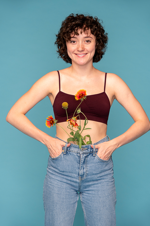 Young pretty cheerful brunette with bunch of wildflowers in blue jeans holding hands in pockets while standing in front of camera