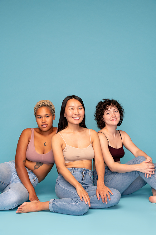 Three young contemporary cheerful women of Asian, Caucasian and African ethnicities sitting on the floor against blue background