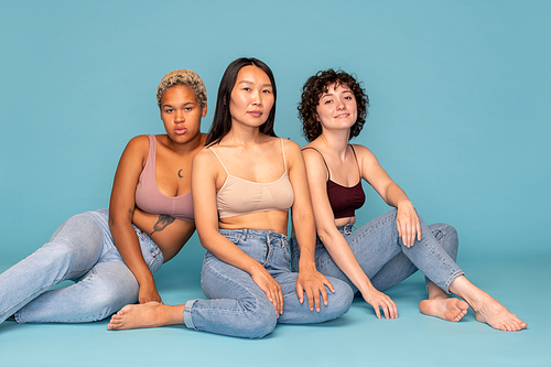 Three young contemporary females of Asian, Caucasian and African ethnicities sitting in front of camera against blue background