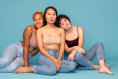 Three young affectionate and friendly women in tanktops and blue jeans sitting close to one another on the floor in front of camera