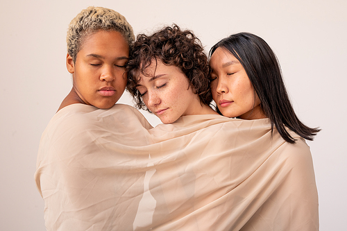 Group of young intercultural females with closed eyes wrapped into white linen while standing close to one another in front of camera