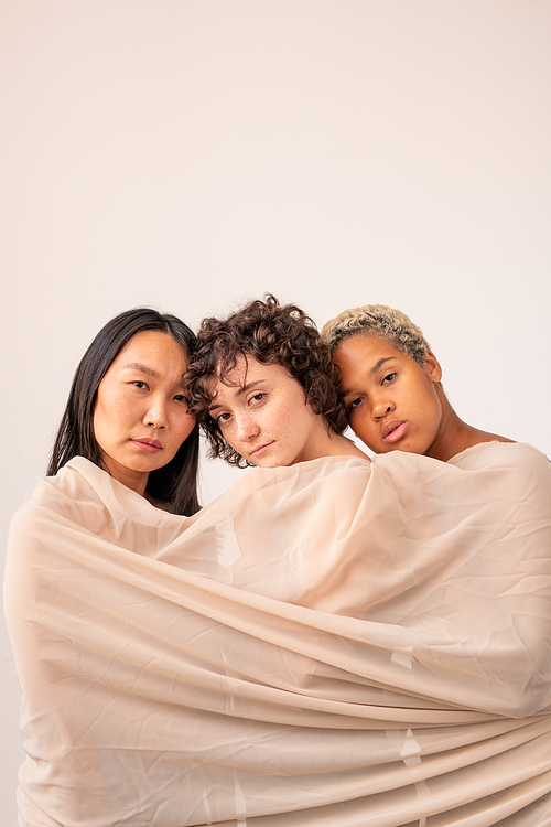 Asian, Caucasian and African girls wrapped into white linen standing close to one another in front of camera in isolation and looking at you