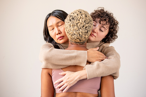 Two young females of Caucasian and Asian ethnicities in one sweater embracing African woman with short dyed blond hair in isolation