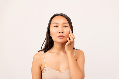Korean or Chinese girl with dark long hair having undereye mask while taking care of her skin in front of camera over white background