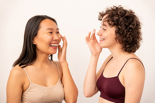 Two young laughing intercultural females in tanktops applying rejuvenating cream on undereye area while looking at one another in isolation