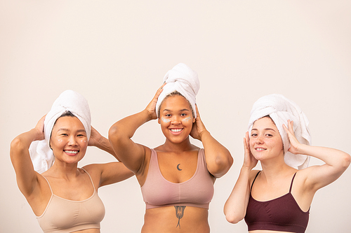 Young happy Asian, African and Caucasian women with white soft towels on heads and tanktops standing in front of camera after bath