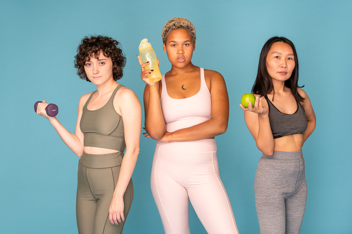 Three young pretty active females of various ethnicities in tracksuits standing in row in front of camera against blue background