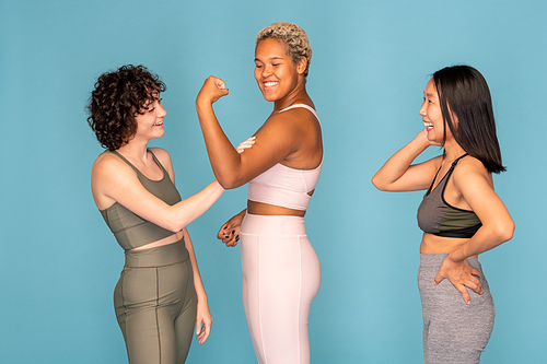 Happy young African woman showing strength while Caucasian girl in tracksuit touching her muscles and Asian female laughing at them