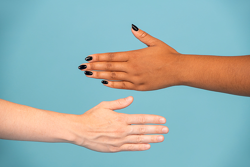 Hands of young women of Caucasian and African ethnicities with white and dark skin stretching towards each other against blue background