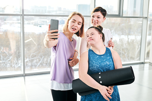 Happy young fitness instructor and two active females looking at smartphone camera while making selfie after workout