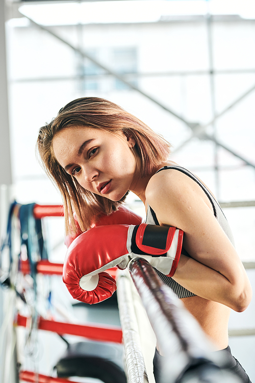Pretty young blond sportswoman in red boxing gloves looking at you while leaning against bars of ring after training in gym