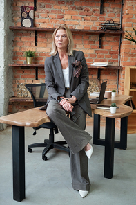 Pensive serious mature business lady in fashionable suit sitting on table in loft office