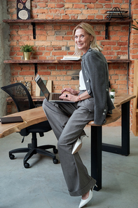 Portrait of cheerful successful mature businesswoman sitting on table and using modern laptop in office