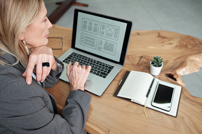 Over shoulder view of serious pensive businesswoman sitting at table and using laptop while working on ui design