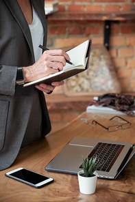 Close-up of unrecognizable businesswoman in jacket standing at desk and writing down ideas in diary