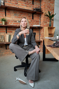 Contemplative mature businesswoman in gray suit keeping bare foot on other leg while drinking coffee in own office