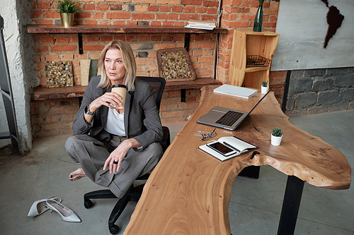 Pensive mature business lady sitting with crossed legs on office chair and drinking coffee while brainstorming about project