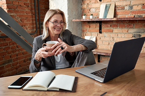 Smiling mature businesswoman drinking coffee and using laptop while communicating with colleague online