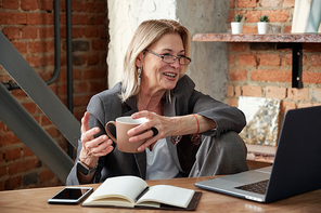 Positive mature businesswoman in glasses sitting in own office and drinking coffee while communicating with colleague via laptop app